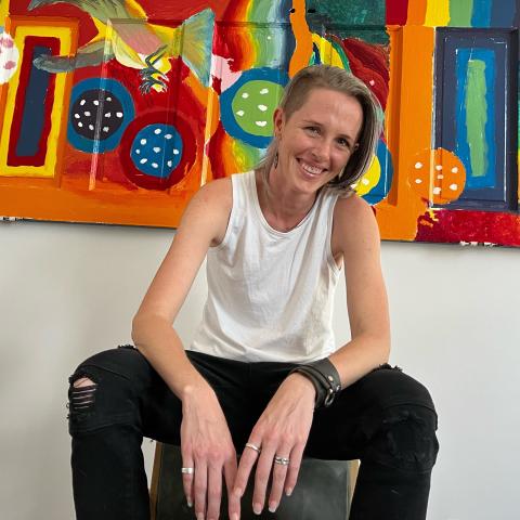 A blond white person with medium length hair with an undercut, a nose piercing, and wearing a white tank top and black pants with a hole on the right knee. Their hands are casually resting in their knees and they are smiling. 