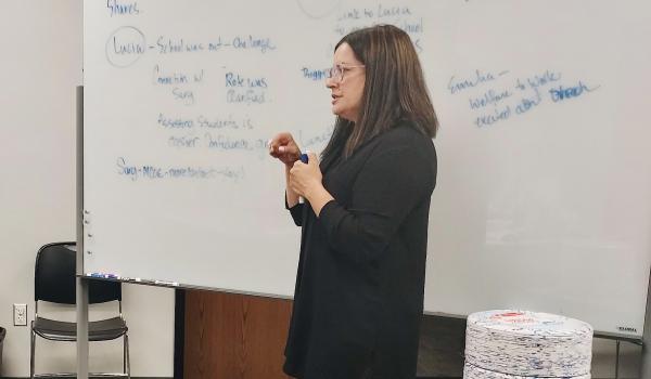 A woman dressed in black with dark brown hair wearing glasses presenting in front of a white board with writing on it.